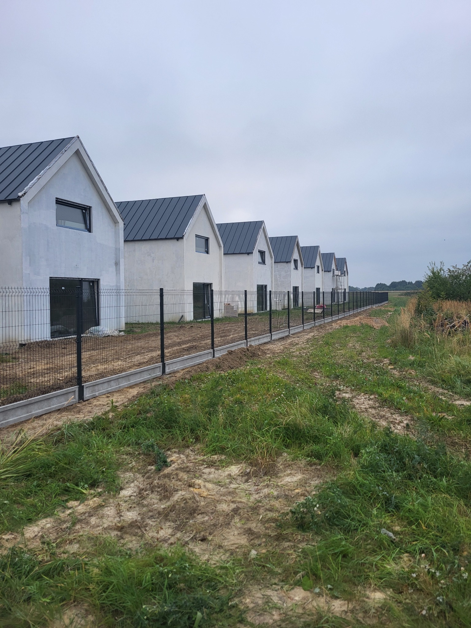 Długa linia ogrodzenia panelowego z podmurówką, oddzielająca szereg nowych domów od łąki. Ciemne panele kontrastują z jasnymi elewacjami budynków.