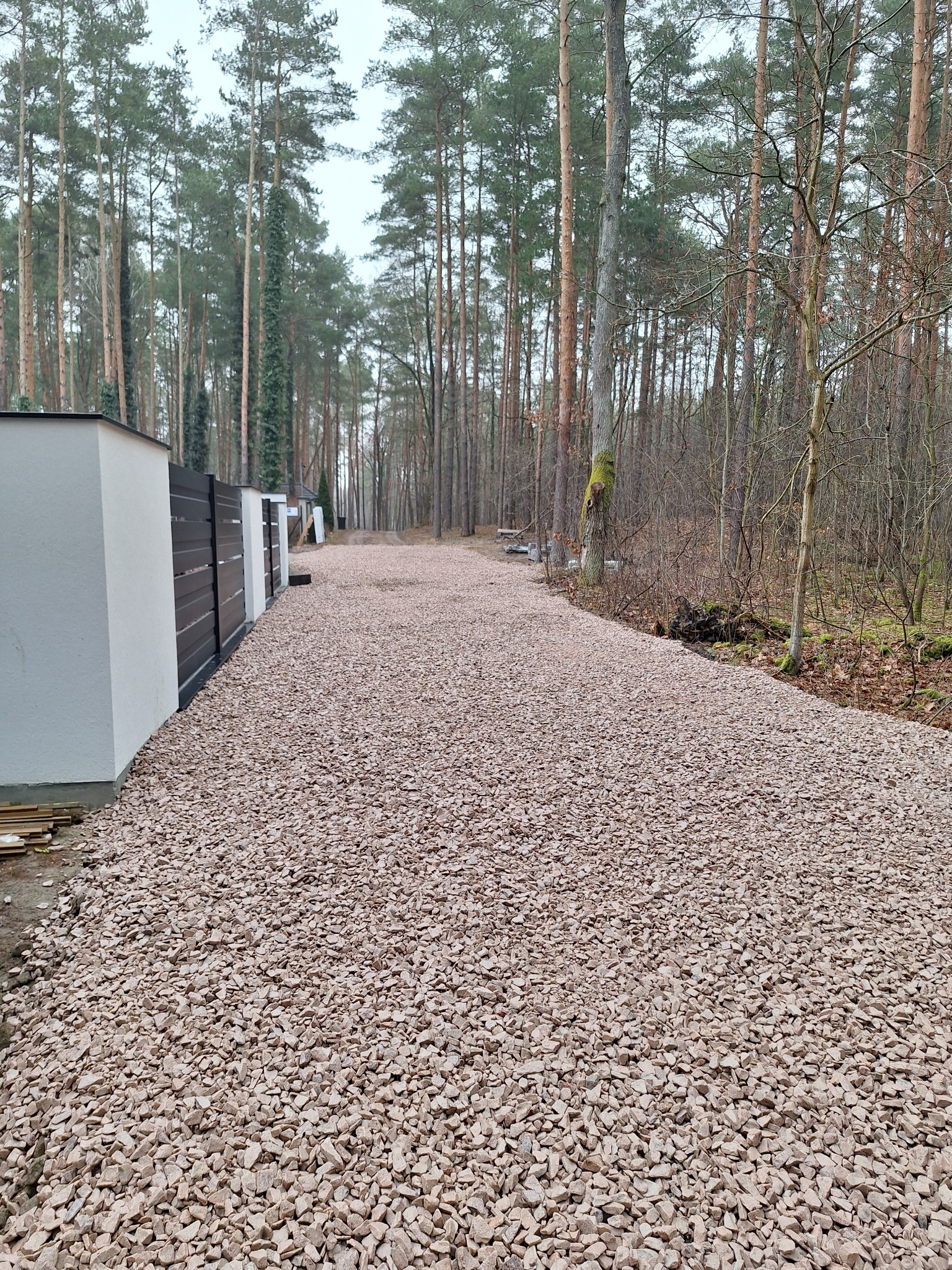 Droga wysypana żwirem prowadząca przez las, obok nowoczesnego ogrodzenia z białymi słupkami i czarnymi panelami. Widok perspektywiczny, pochmurna pogoda.