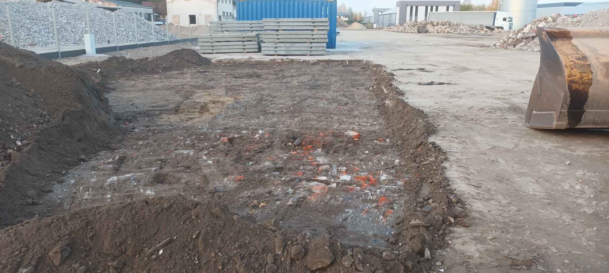 Wykop w ziemi z widocznymi śladami opon, na tle składowiska gruzu i zaparkowanego sprzętu budowlanego. W tle widoczne budynki przemysłowe i niebieski kontener.