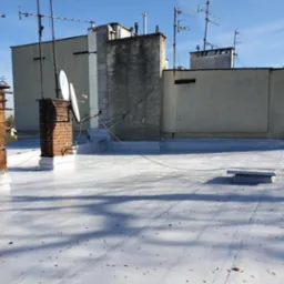 Biała, świeżo pokryta powierzchnia dachu budynku z ceglanym kominem i antenami satelitarnymi na tle błękitnego nieba. Widoczne detale wykończenia i faktura nowej powłoki dachowej.