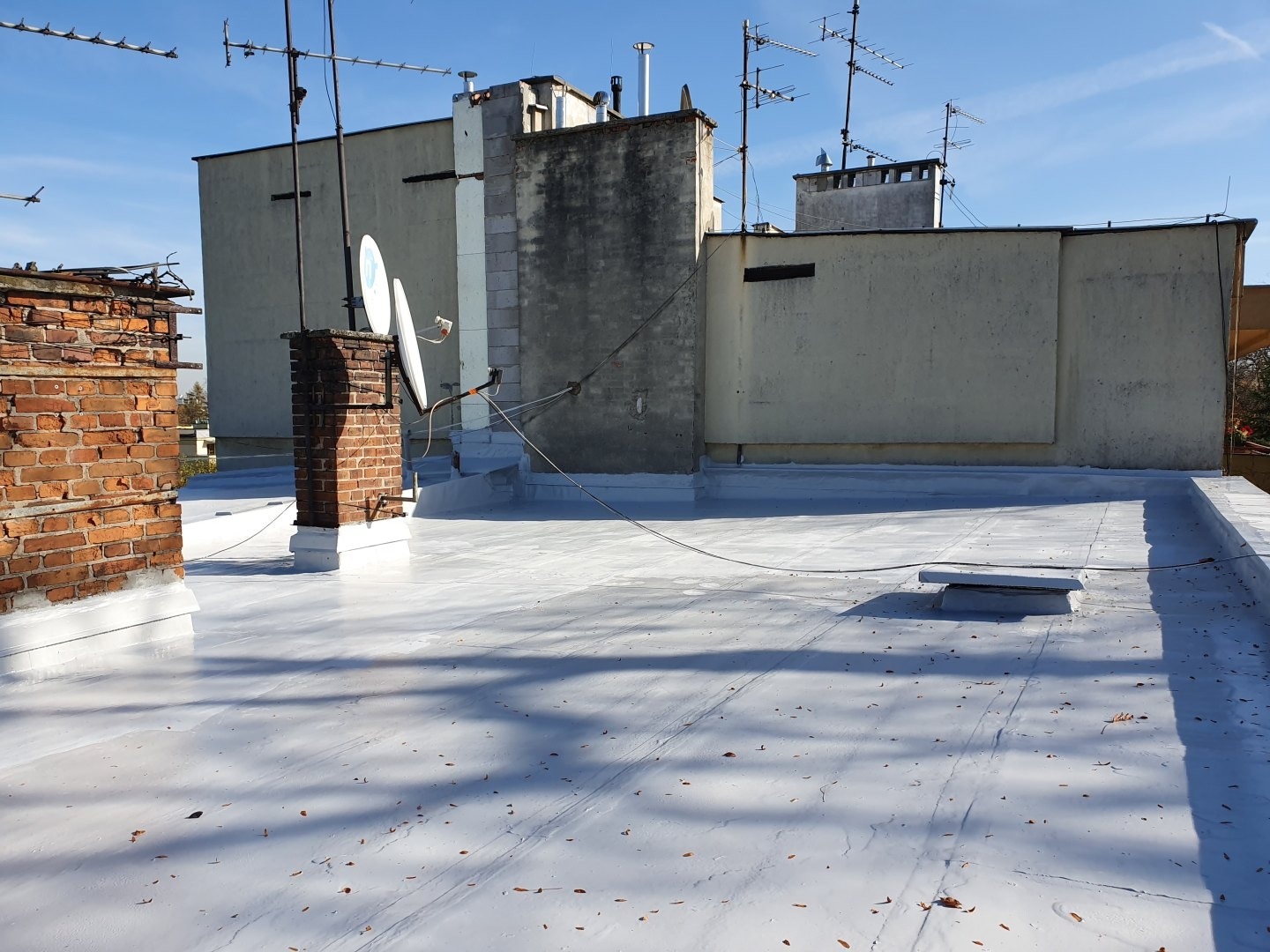 Biała, świeżo pokryta powierzchnia dachu budynku z ceglanym kominem i antenami satelitarnymi na tle błękitnego nieba. Widoczne detale wykończenia i faktura nowej powłoki dachowej.