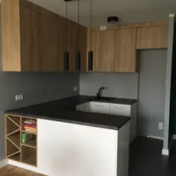 Nowoczesna kuchnia z drewnianymi szafkami wiszącymi, czarnym blatem i białym wykończeniem wyspy kuchennej z półkami na książki.