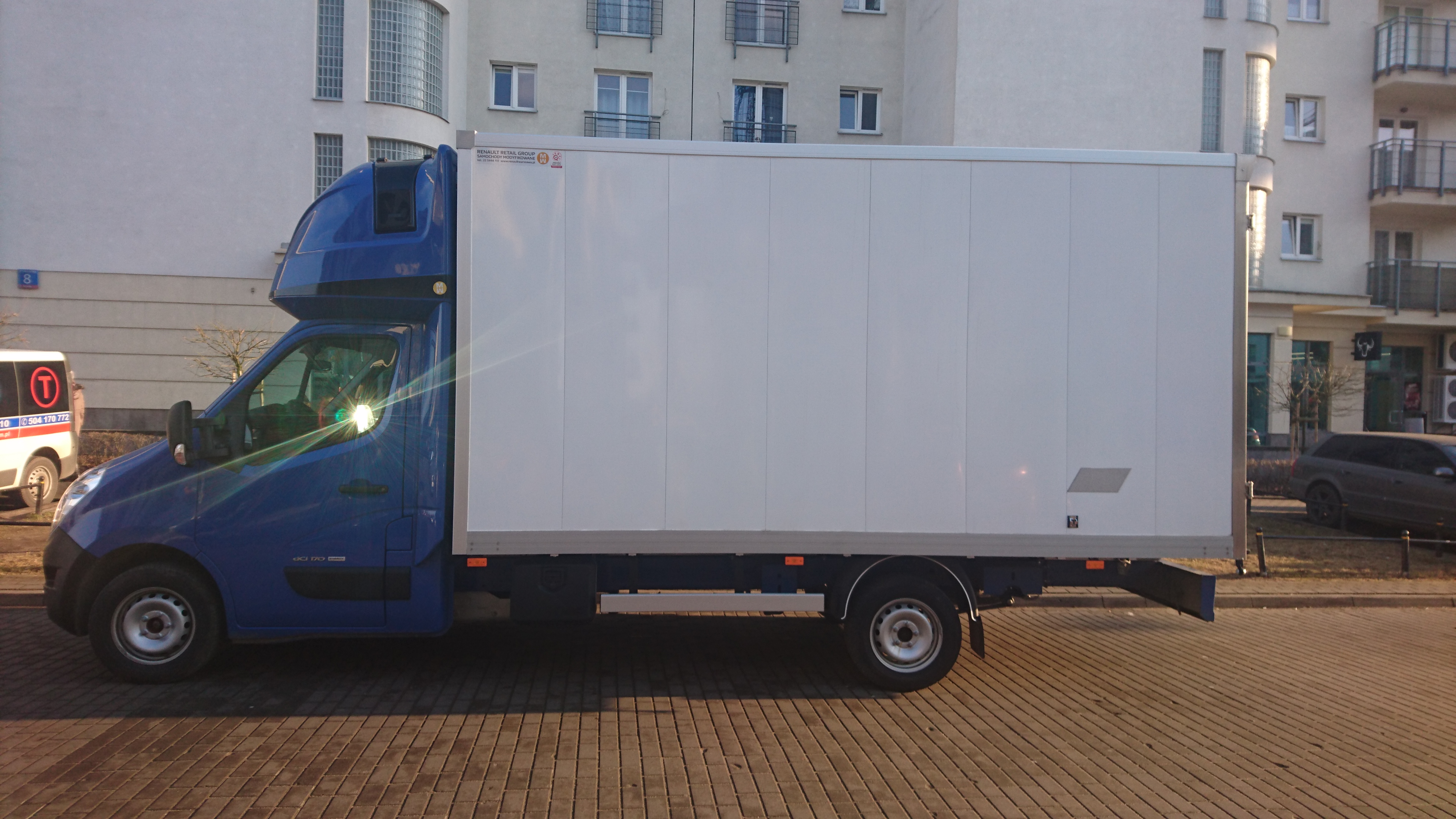 Niebieski samochód dostawczy z białą zabudową kontenerową zaparkowany na brukowanej ulicy przed budynkiem mieszkalnym.