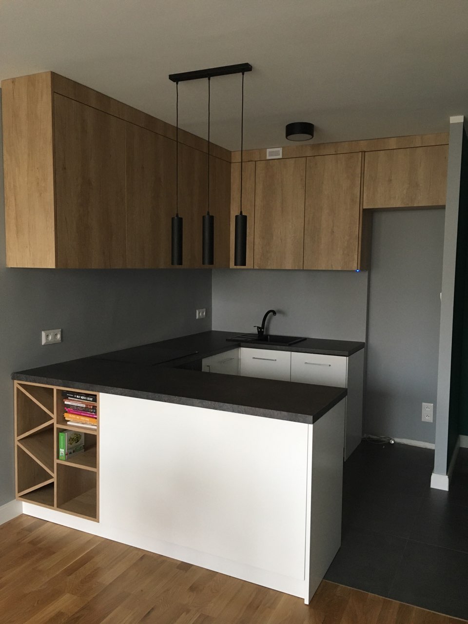 Nowoczesna kuchnia z drewnianymi szafkami wiszącymi, czarnym blatem i białym wykończeniem wyspy kuchennej z półkami na książki.