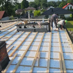 Dach w trakcie ocieplania z widocznymi kominami i pracownikami. Biała termoizolacja pokryta drewnianą konstrukcją. Budynki mieszkalne w tle.