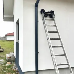 Zamontowana, grafitowa rynna spustowa na jasnej elewacji domu jednorodzinnego. Przy ścianie oparta drabina. Widoczne fragmenty dachu z dachówką.
