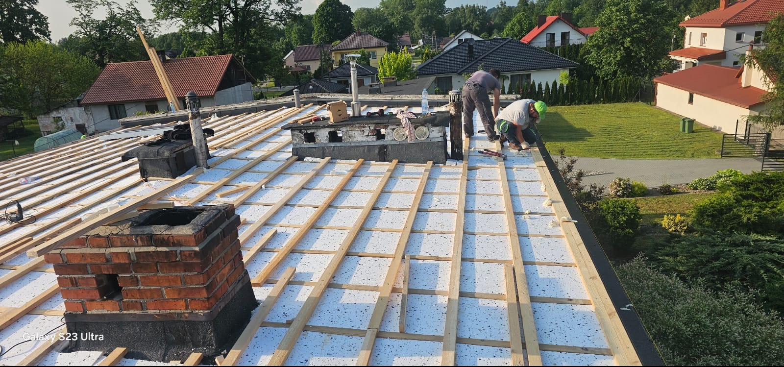 Dach w trakcie ocieplania z widocznymi kominami i pracownikami. Biała termoizolacja pokryta drewnianą konstrukcją. Budynki mieszkalne w tle.
