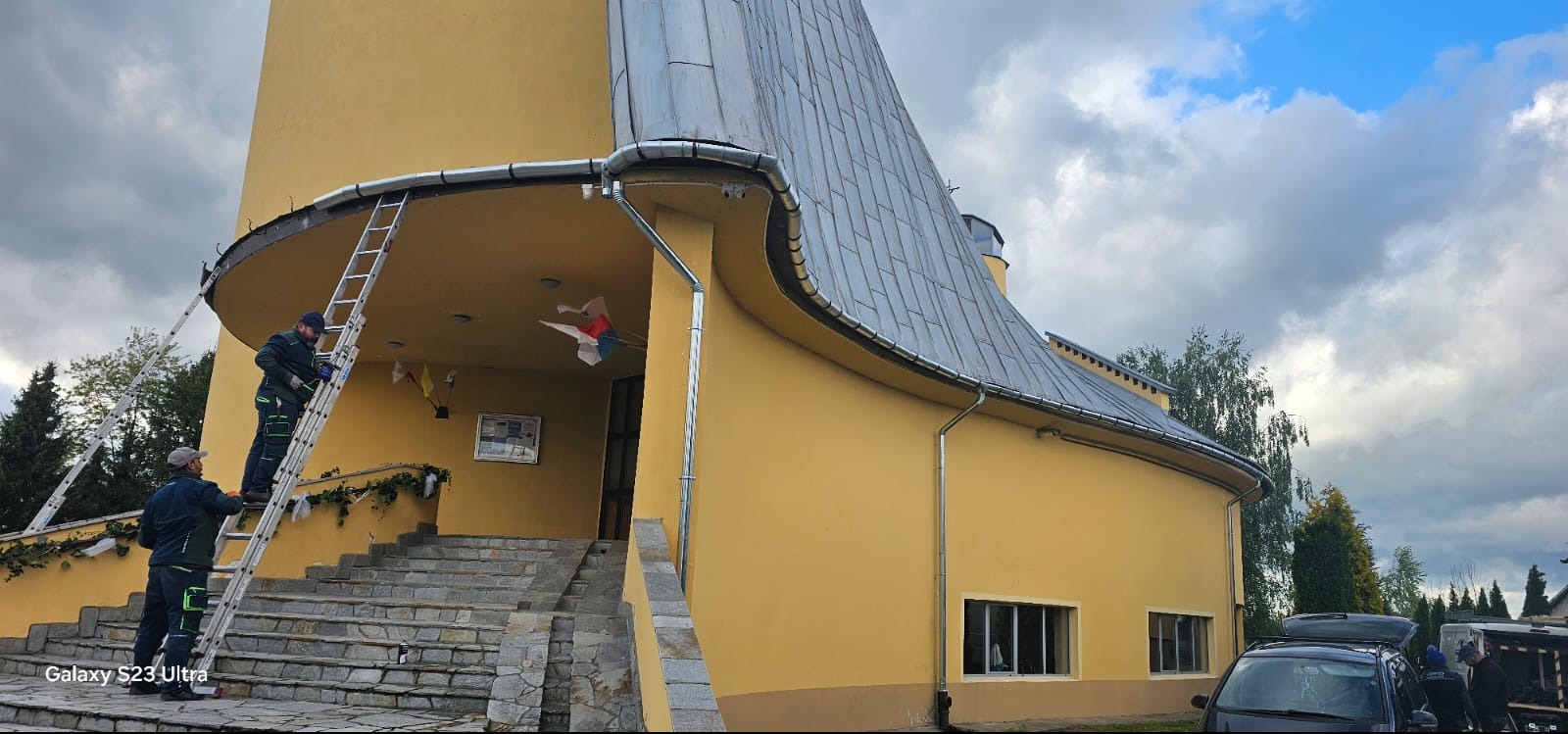 Pracownicy na drabinach montują rynny na nietypowym, żółtym budynku z szarym dachem. Widoczne elementy obróbki blacharskiej i charakterystyczna architektura budynku.