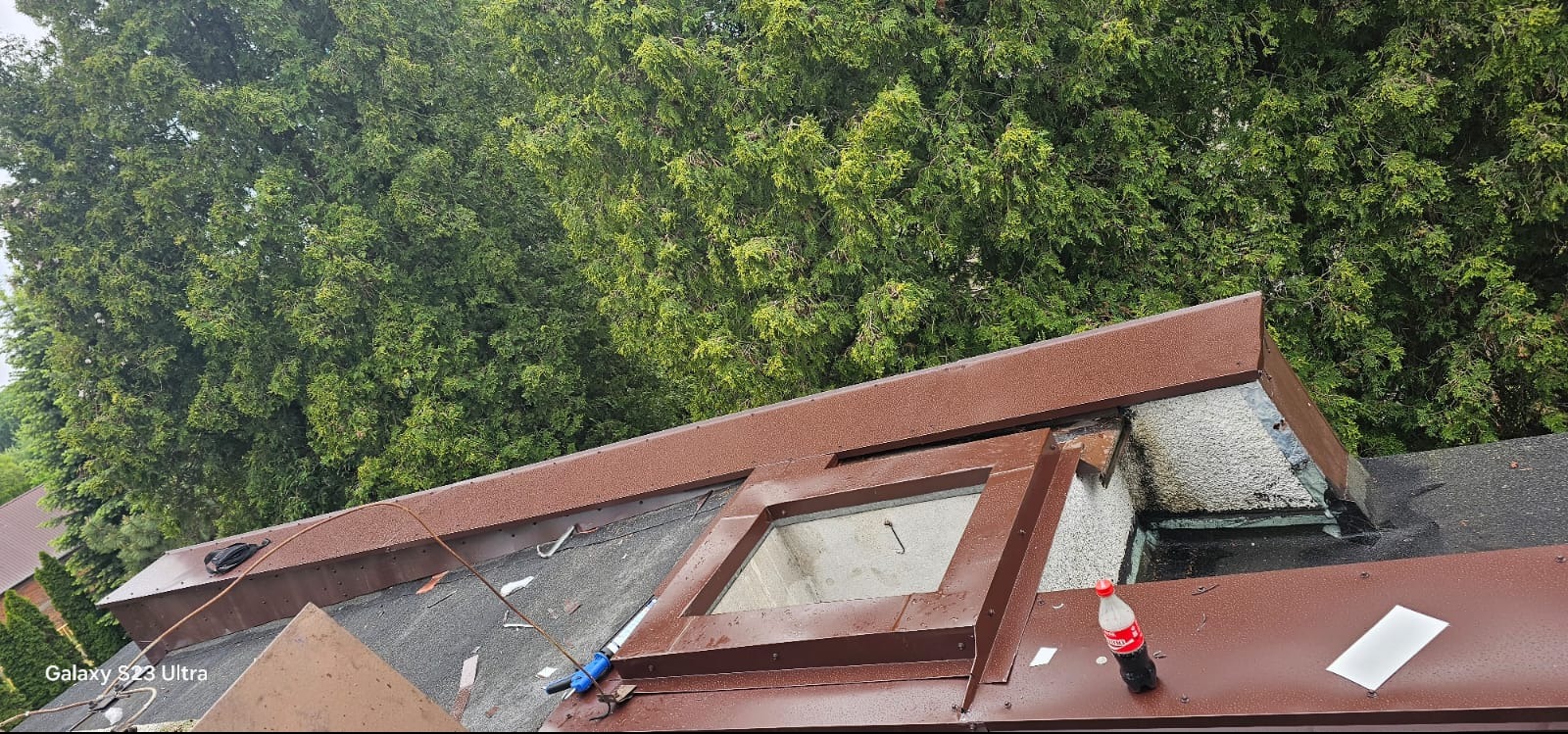 Fragment dachu z brązowymi obróbkami blacharskimi i oknem dachowym w trakcie prac. W tle drzewa. Widoczne narzędzia i butelka napoju.