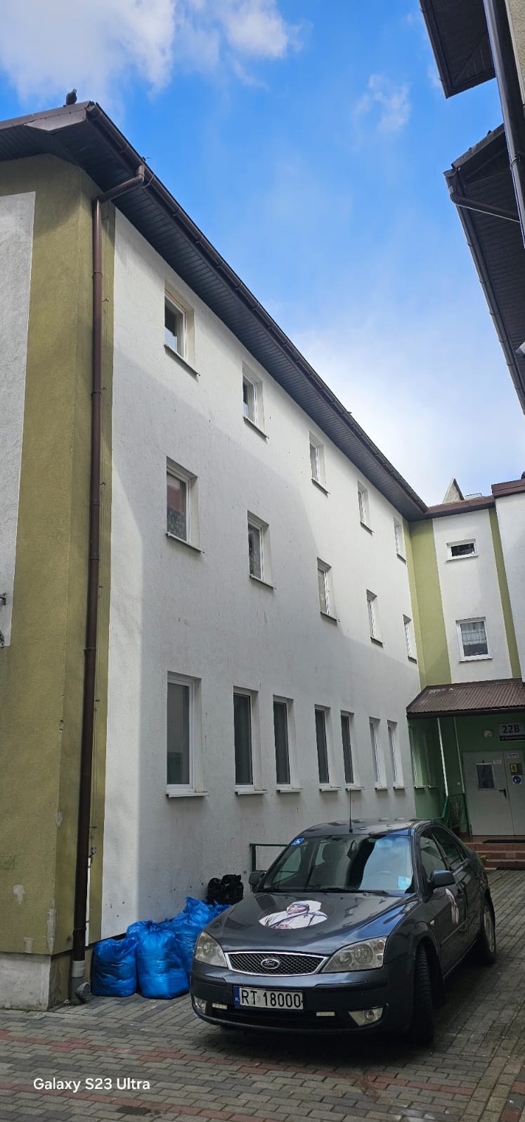 Elewacja budynku z rynnami i dachem widoczna z dołu, z zaparkowanym autem i workami. Widoczny fragment nieba z chmurami. Ujęcie z dołu.