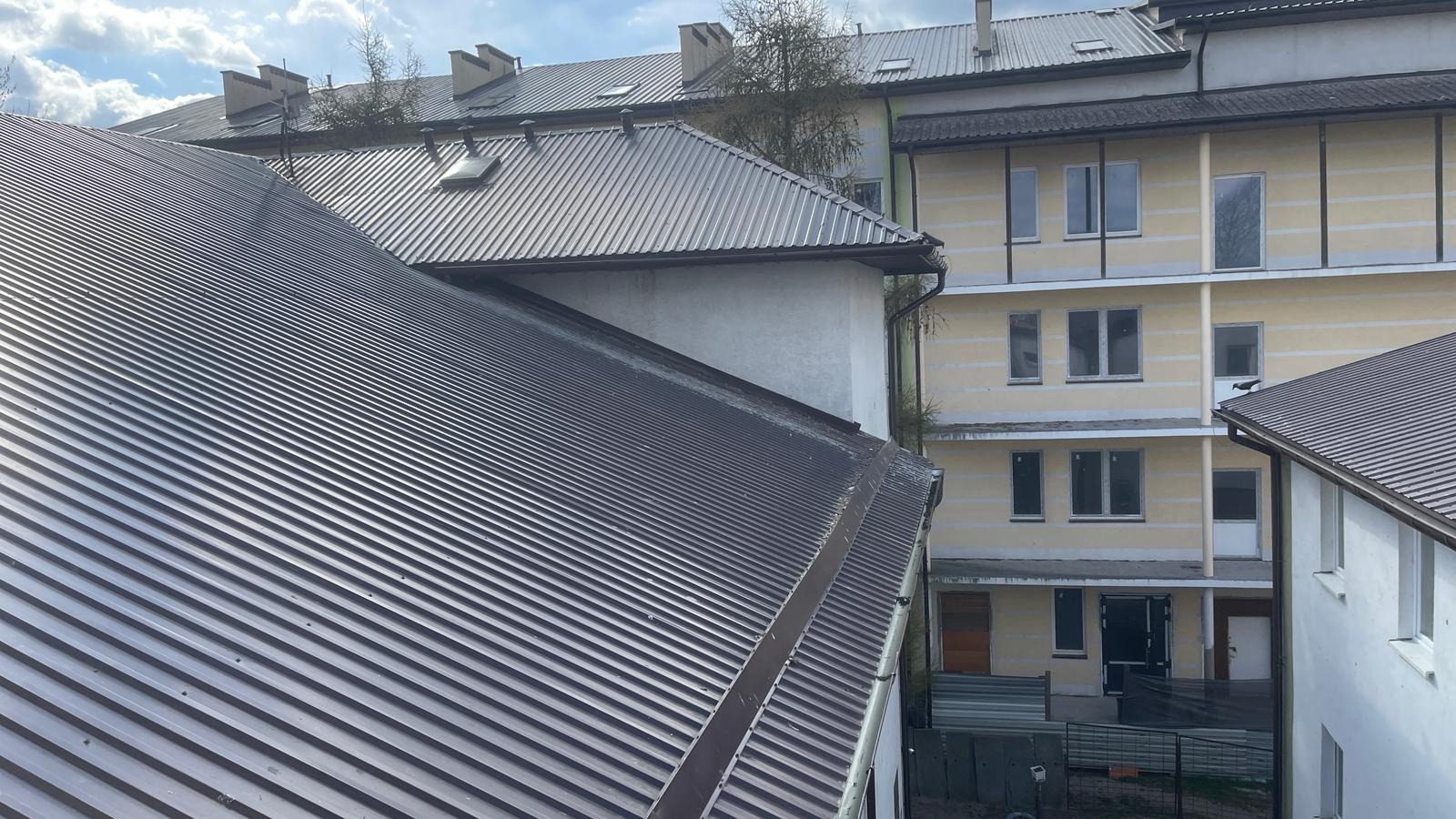 Szare, trapezowe dachy budynków mieszkalnych widziane z góry, z widocznymi rynnami i elementami obróbki blacharskiej na tle błękitnego nieba z chmurami.