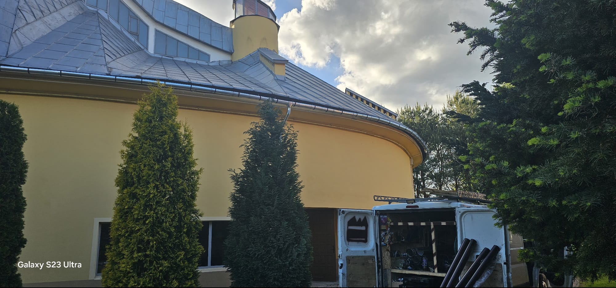 Elewacja budynku z szarą blachą dachową i rynnami, widoczny otwarty bus z narzędziami. Uwagę przykuwa zaokrąglona ściana budynku i iglaki.