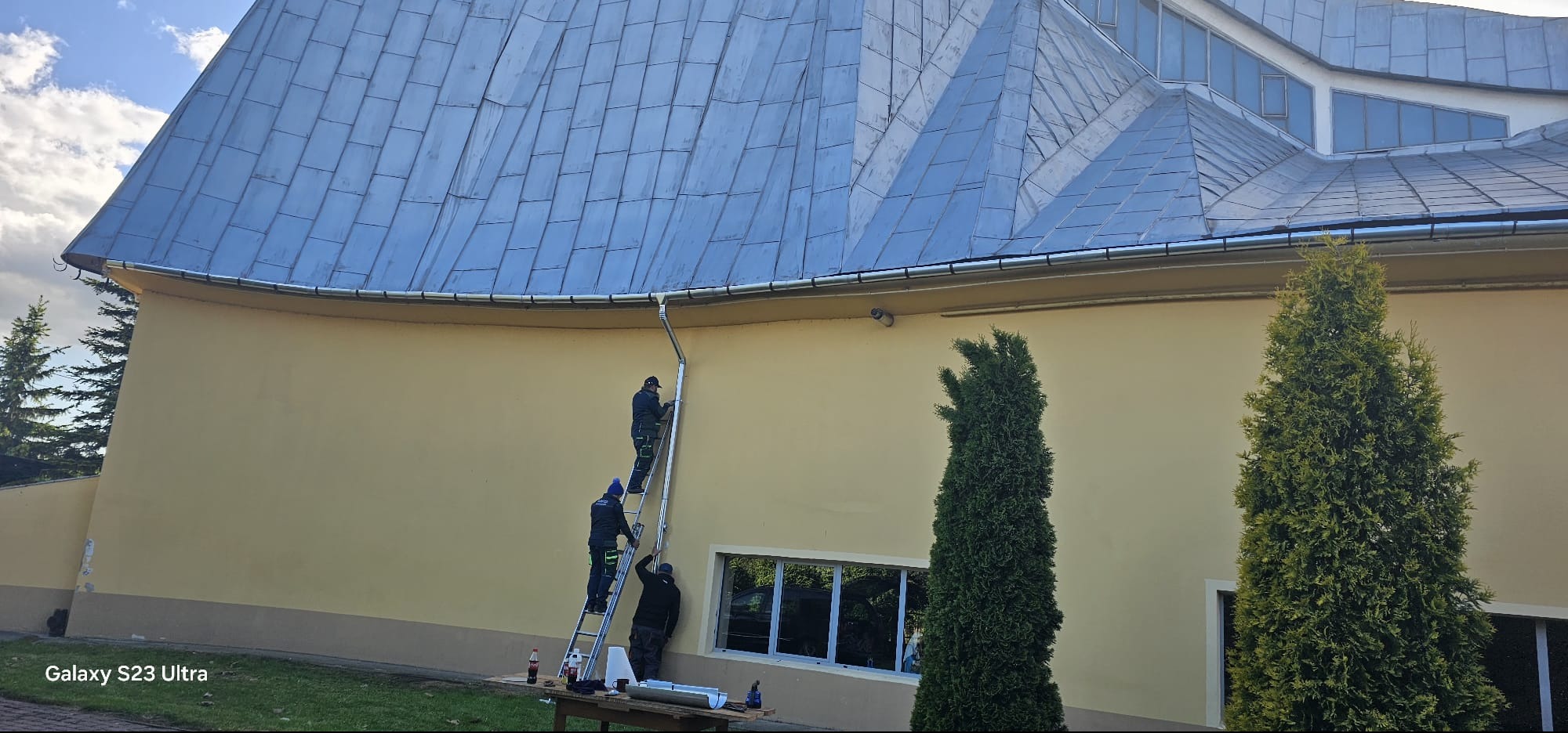 Pracownicy na drabinie montują rynnę spustową na budynku z blachodachówką trapezową, widoczne narzędzia i materiały na stole roboczym.