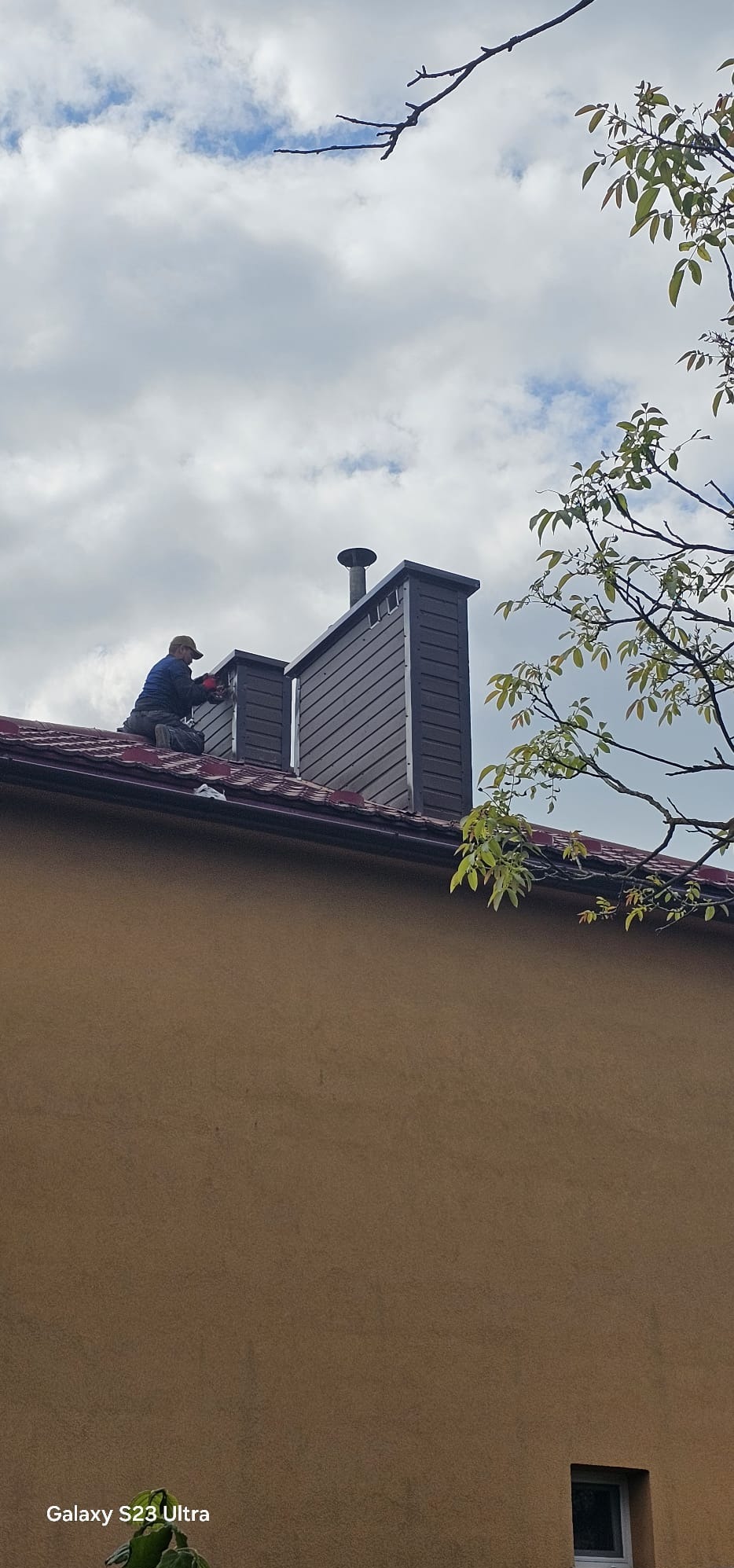 Praca na dachu w Rzeszowie: dekarz montuje obróbki blacharskie komina z brązowej blachy na tle pochmurnego nieba i zieleni drzew.