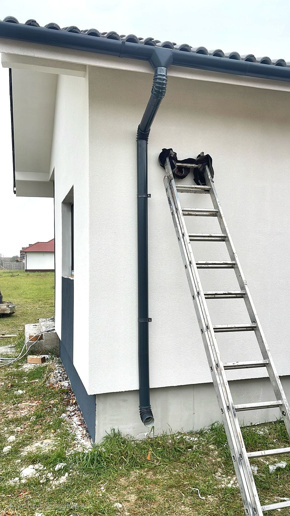 Zamontowana, grafitowa rynna spustowa na jasnej elewacji domu jednorodzinnego. Przy ścianie oparta drabina. Widoczne fragmenty dachu z dachówką.