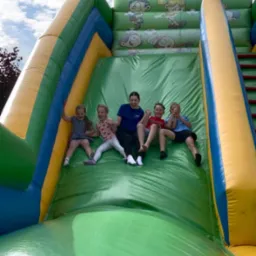 Radosne dzieci z animatorką zjeżdżają na dmuchanej, kolorowej zjeżdżalni. Zabawa w plenerze w słoneczny dzień. Uśmiechy i pozytywna energia.