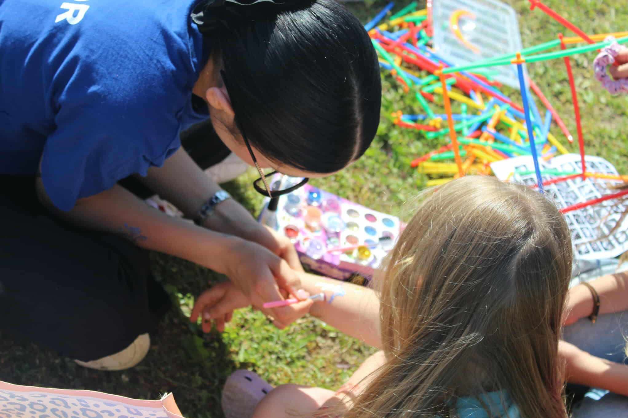 Animator maluje wzór na ramieniu dziewczynki w plenerze. Kolorowe farby i akcesoria leżą obok, tworząc radosną atmosferę zabawy na trawie.