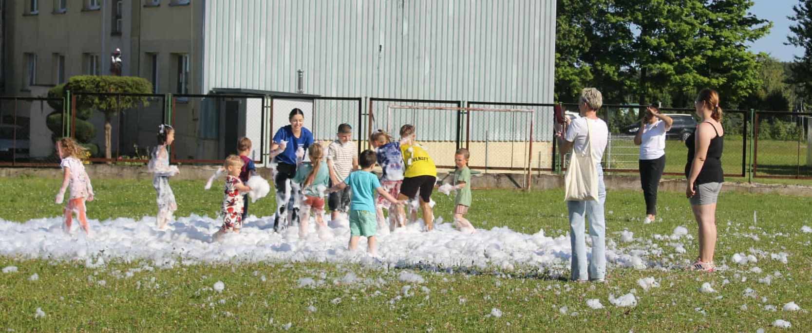 Animator w akcji: grupa dzieci bawi się w pianie na zielonym trawniku, a osoby dorosłe robią zdjęcia. W tle budynek i drzewa.