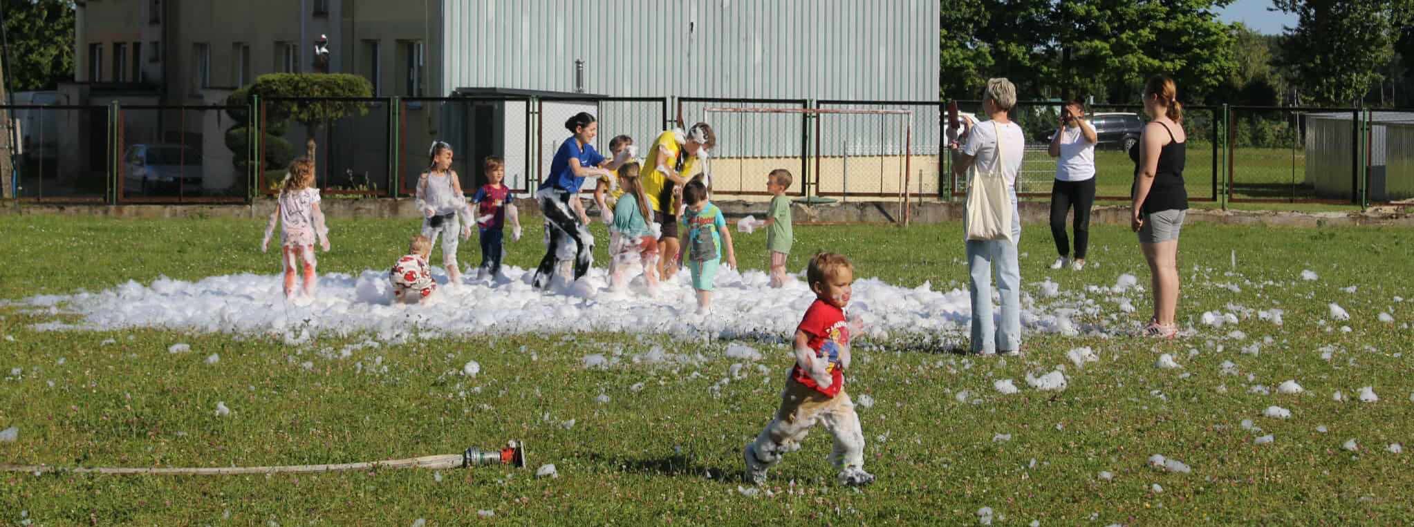 Radosne dzieci w pianie na trawie, zabawa z animatorem w Gdańsku. Rodzice robią zdjęcia. W tle budynek i ogrodzenie.