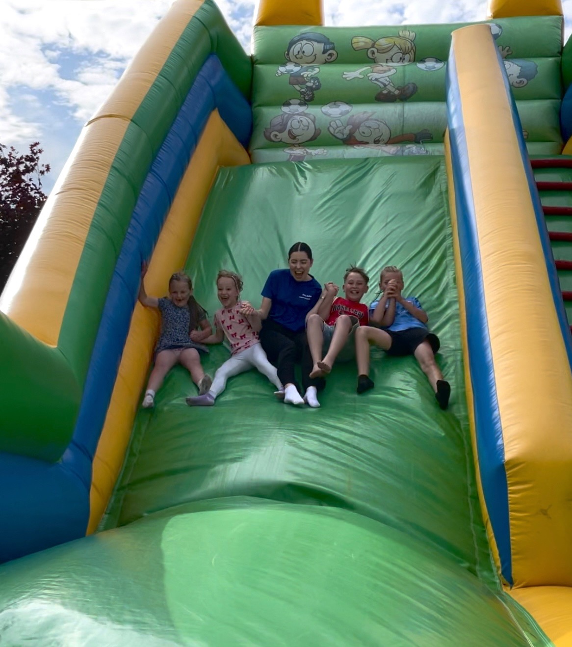 Radosne dzieci z animatorką zjeżdżają na dmuchanej, kolorowej zjeżdżalni. Zabawa w plenerze w słoneczny dzień. Uśmiechy i pozytywna energia.