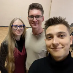 Trzy uśmiechnięte osoby, w tym potencjalny instruktor z przodu, pozują do selfie w sali z drewnianą podłogą, sugerując atmosferę szkoły tańca.