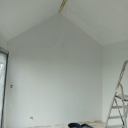 Kwiecień Krystian Usługi remontowo budowlane - Wnętrze w trakcie malowania: białe ściany, drabina, wiaderka z farbą i wałki malarskie. Widoczne niedokończone prace przy suficie i wystające przewody.
