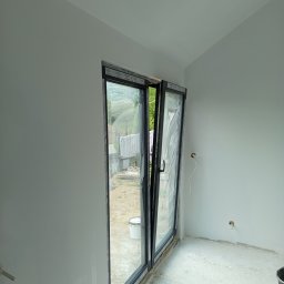 Kwiecień Krystian Usługi remontowo budowlane - Wnętrze po malowaniu ścian na biało, z otwartym oknem balkonowym i widocznymi puszkami elektrycznymi. Świeżo pomalowane ściany i sufit. Widok na zewnątrz.