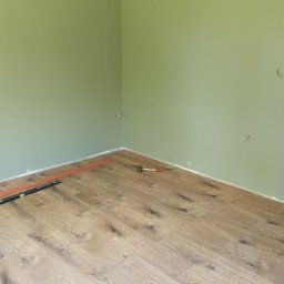 Kwiecień Krystian Usługi remontowo budowlane - Pokój z zielonymi ścianami i częściowo ułożoną podłogą z paneli. Widoczne narzędzia: poziomica i młotek. W tle wystające kable elektryczne.