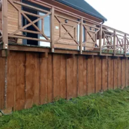 Drewniany budynek z tarasem, widok z dołu. Brązowe deski elewacyjne, zielona trawa. Charakterystyczne krzyżowe wzory na balustradzie tarasu.