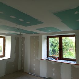 Kwiecień Krystian Usługi remontowo budowlane - Wnętrze w trakcie prac w Gorzanowie: ściany przygotowane do malowania, częściowo położona płyta karton-gips na suficie z widocznymi łączeniami. Dwa okna z drewnianą stolarką.