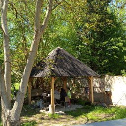 Kwiecień Krystian Usługi remontowo budowlane - Drewniana altana z dachem krytym gontem, otoczona zielenią drzew. Wewnątrz siedzą osoby przy stole. Na zewnątrz widoczny grill i elementy ogrodzenia.