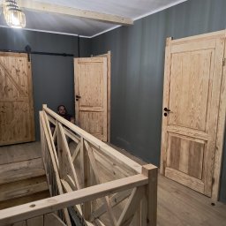 Kwiecień Krystian Usługi remontowo budowlane - Wnętrze z drewnianymi schodami, balustradą i drzwiami z surowego drewna. Mężczyzna siedzi na schodach. Szare ściany, drewniana podłoga. Widoczna lampa wisząca.