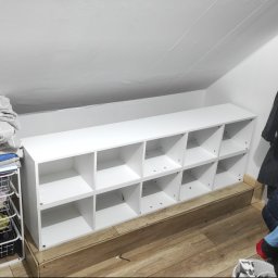 Kwiecień Krystian Usługi remontowo budowlane - Biały regał z przegródkami na poddaszu, obok kosz z tekstyliami i wieszak z ubraniami. Wykończenie wnętrza ze skosami, drewniana podłoga.