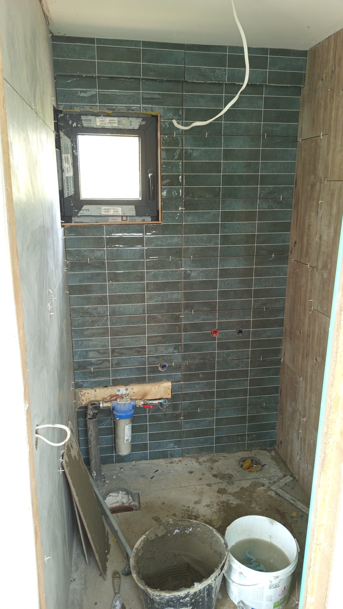 Fragment łazienki w trakcie prac glazurniczych: zielono-szare, wąskie płytki na ścianie z oknem, wiadro z zaprawą i wodą na podłodze. Widoczne krzyżyki dystansowe.