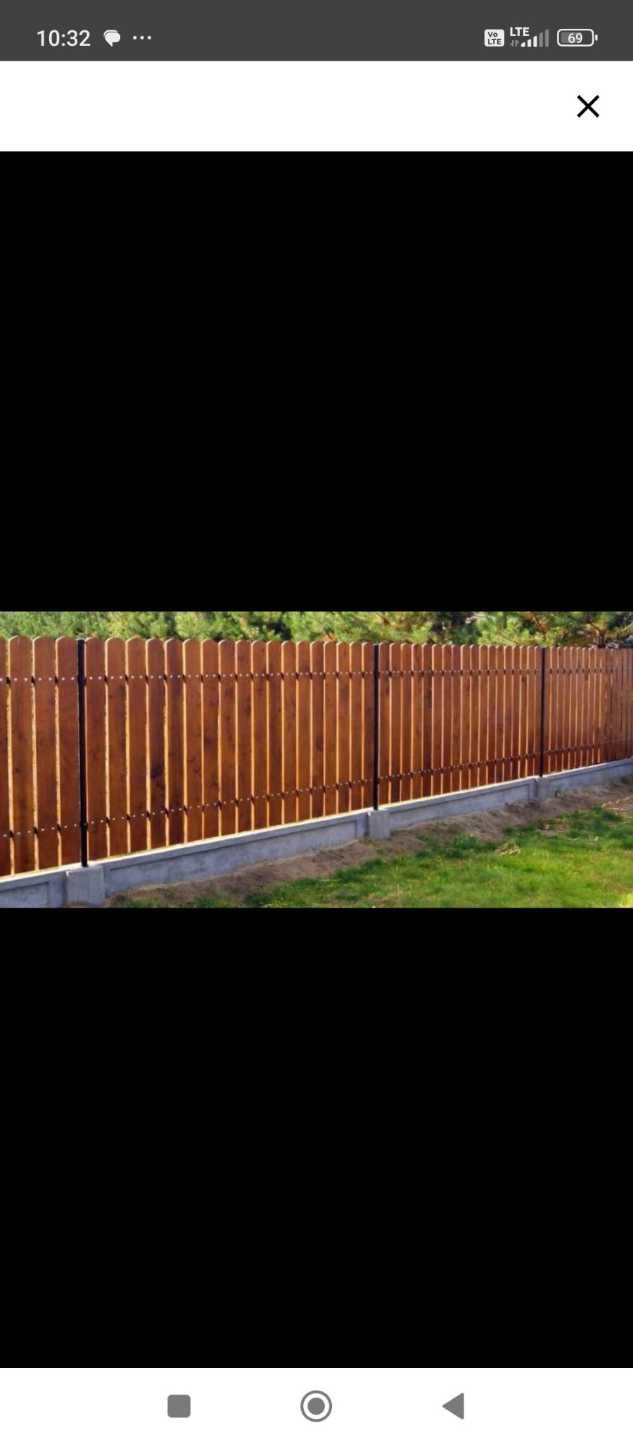 Nowy, brązowy płot drewniany z betonową podmurówką, oddzielający trawnik od zieleni. Słupki metalowe, równomiernie rozmieszczone. Słoneczny dzień.