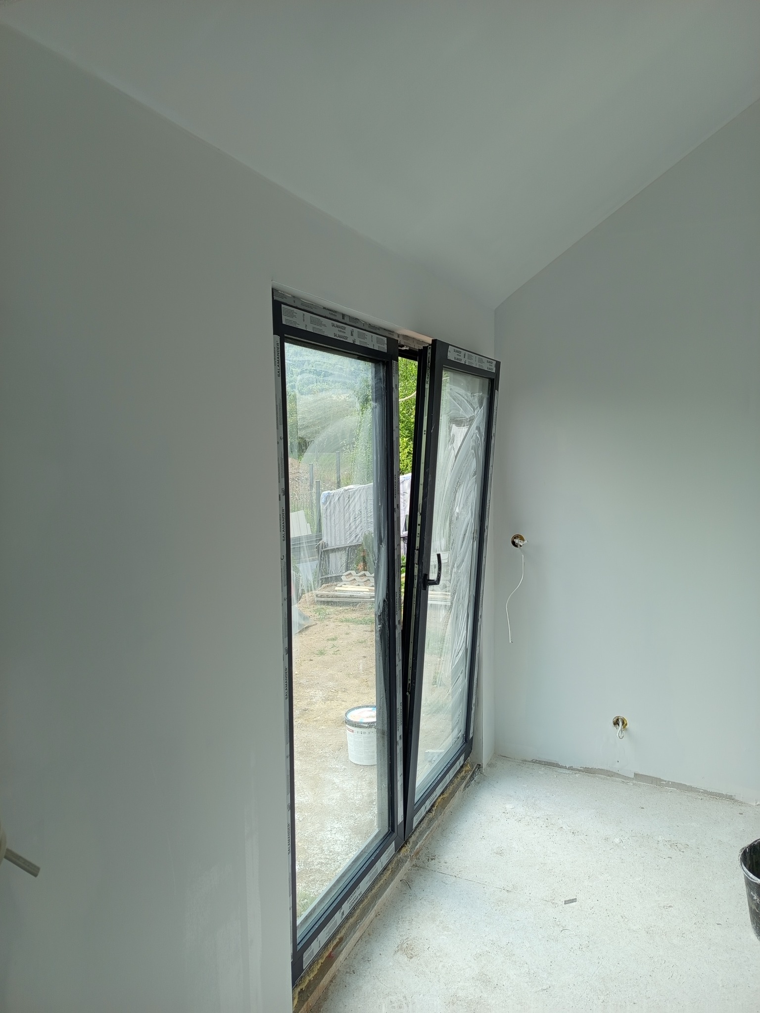 Wnętrze po malowaniu ścian na biało, z otwartym oknem balkonowym i widocznymi puszkami elektrycznymi. Świeżo pomalowane ściany i sufit. Widok na zewnątrz.