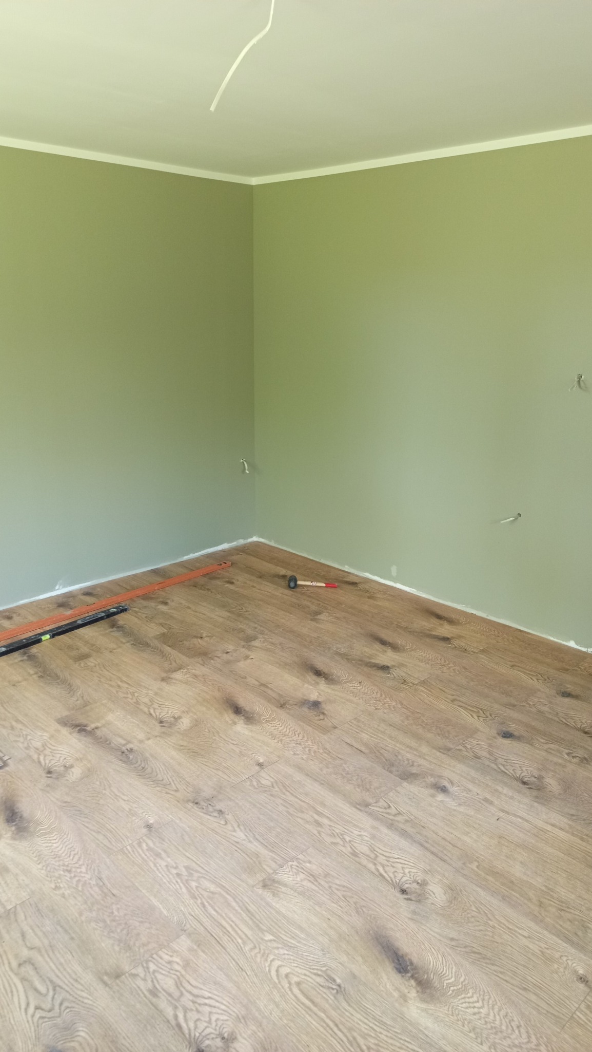 Pokój z zielonymi ścianami i częściowo ułożoną podłogą z paneli. Widoczne narzędzia: poziomica i młotek. W tle wystające kable elektryczne.