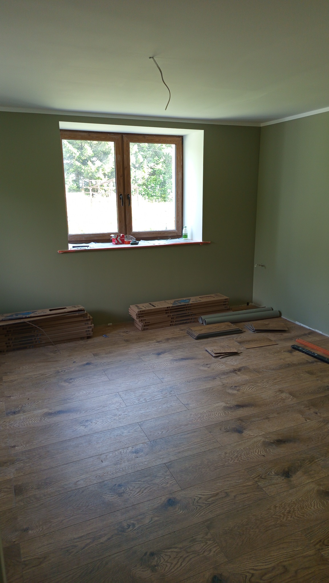 Ułożona podłoga z paneli w pokoju z zielonymi ścianami i oknem. Na podłodze leżą paczki paneli i narzędzia. Widoczny fragment kabla wystającego z sufitu.