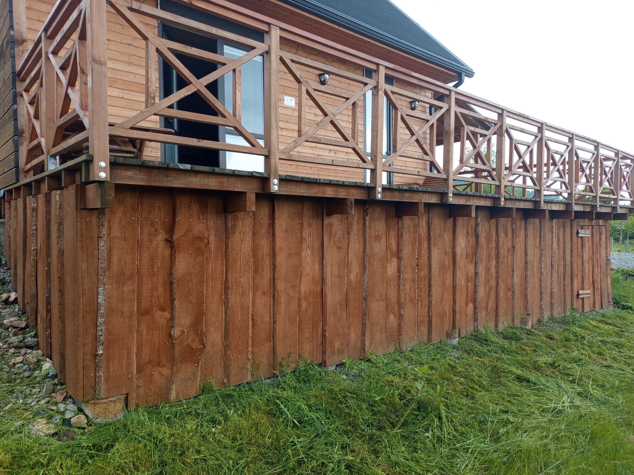 Drewniany budynek z tarasem, widok z dołu. Brązowe deski elewacyjne, zielona trawa. Charakterystyczne krzyżowe wzory na balustradzie tarasu.