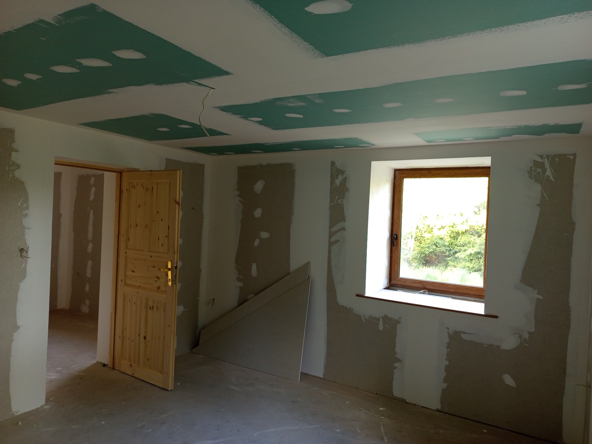 Pokój w trakcie remontu: zielone płyty karton-gips na suficie, ściany częściowo wyszpachlowane, drewniane drzwi, okno z widokiem na zieleń. Widoczne płyty gipsowe.