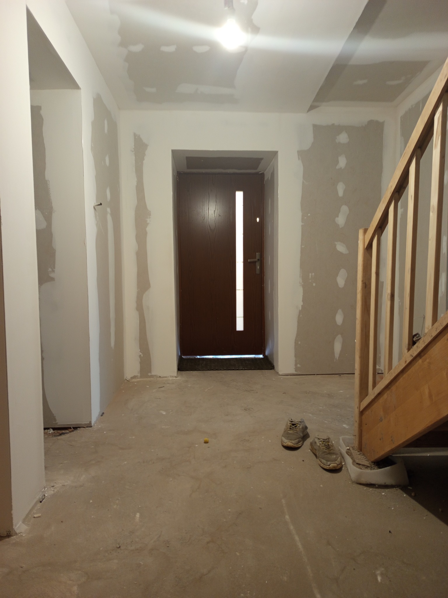 Wnętrze w trakcie remontu: szpachlowane ściany, surowy beton na podłodze, drewniane schody i wejściowe drzwi z przeszkleniem. Dwa buty leżą na podłodze.