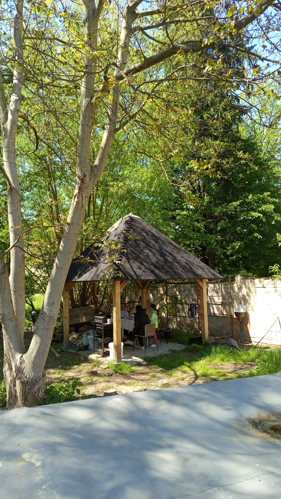 Drewniana altana z dachem krytym gontem, otoczona zielenią drzew. Wewnątrz siedzą osoby przy stole. Na zewnątrz widoczny grill i elementy ogrodzenia.