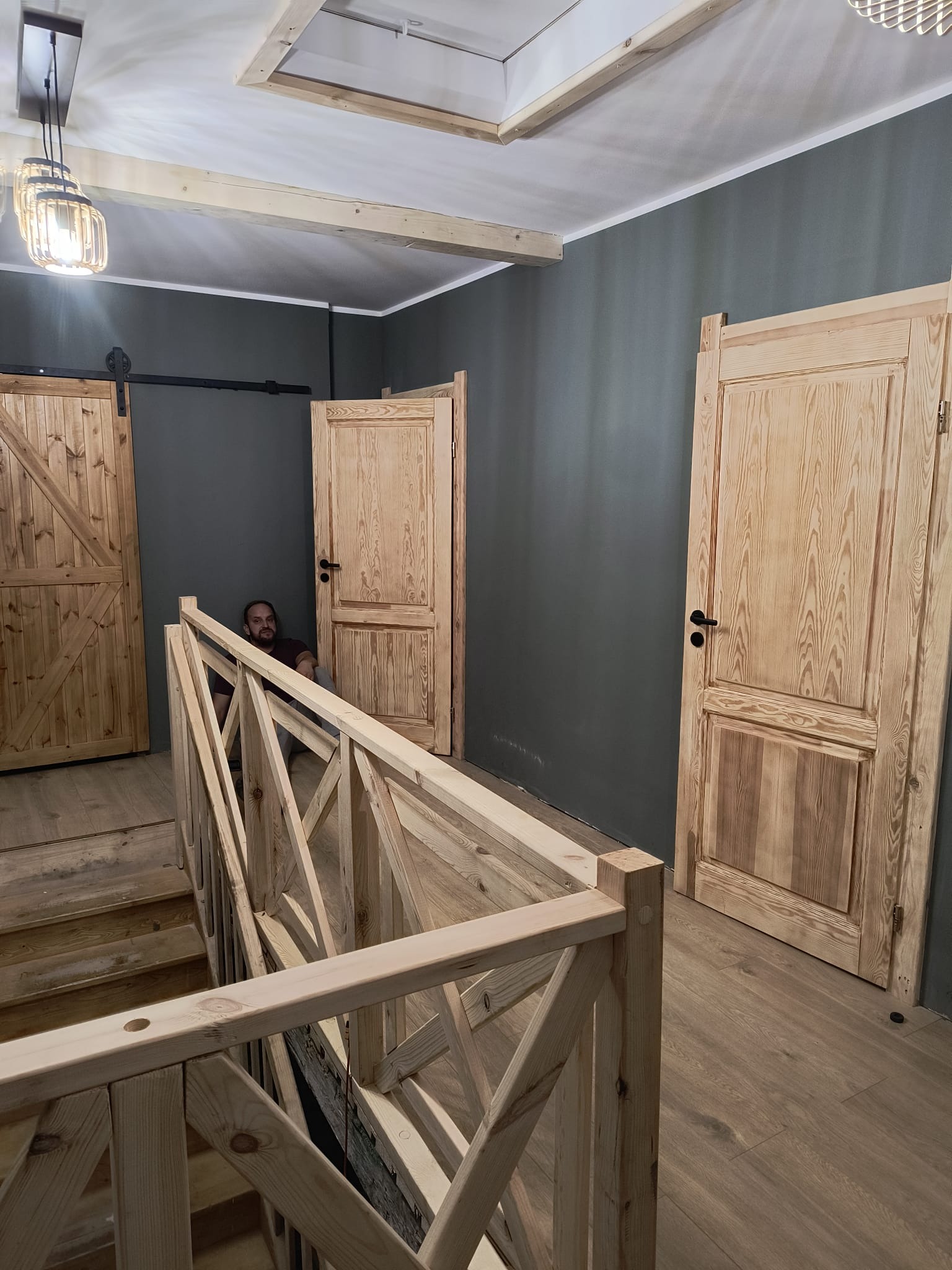 Wnętrze z drewnianymi schodami, balustradą i drzwiami z surowego drewna. Mężczyzna siedzi na schodach. Szare ściany, drewniana podłoga. Widoczna lampa wisząca.