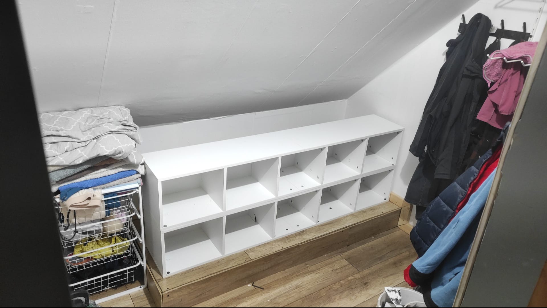 Biały regał z przegródkami na poddaszu, obok kosz z tekstyliami i wieszak z ubraniami. Wykończenie wnętrza ze skosami, drewniana podłoga.