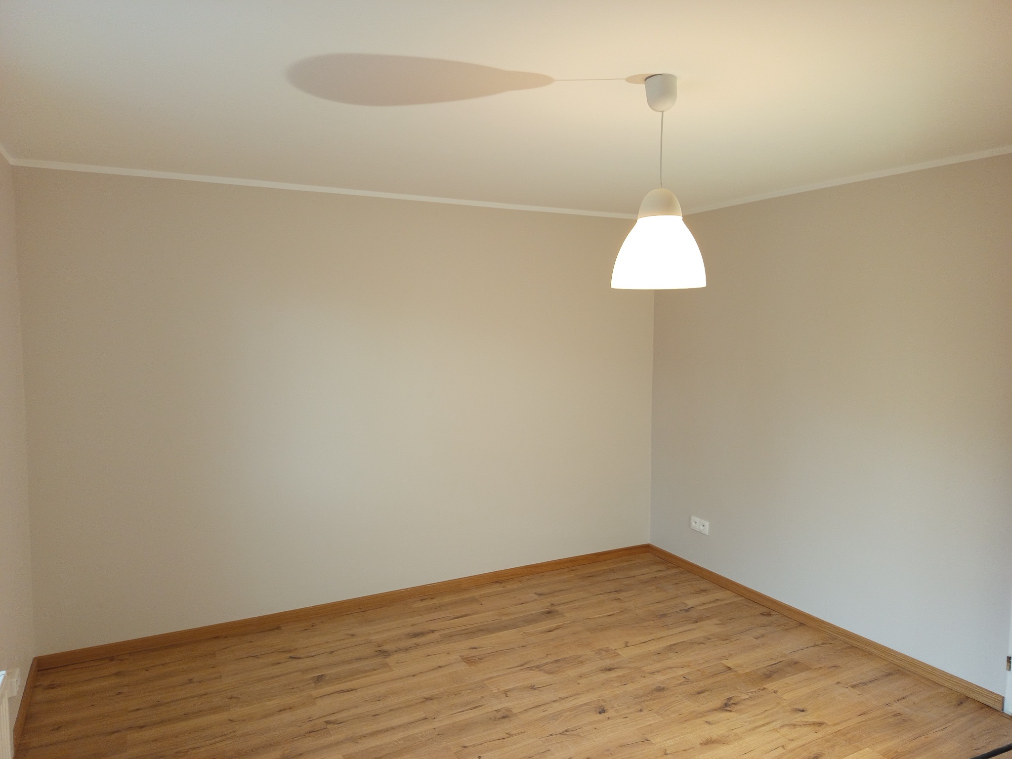 Jasne wnętrze po remoncie: drewnopodobna podłoga, beżowe ściany i nowoczesna lampa wisząca. Widoczny fragment listwy przypodłogowej i gniazdko elektryczne.