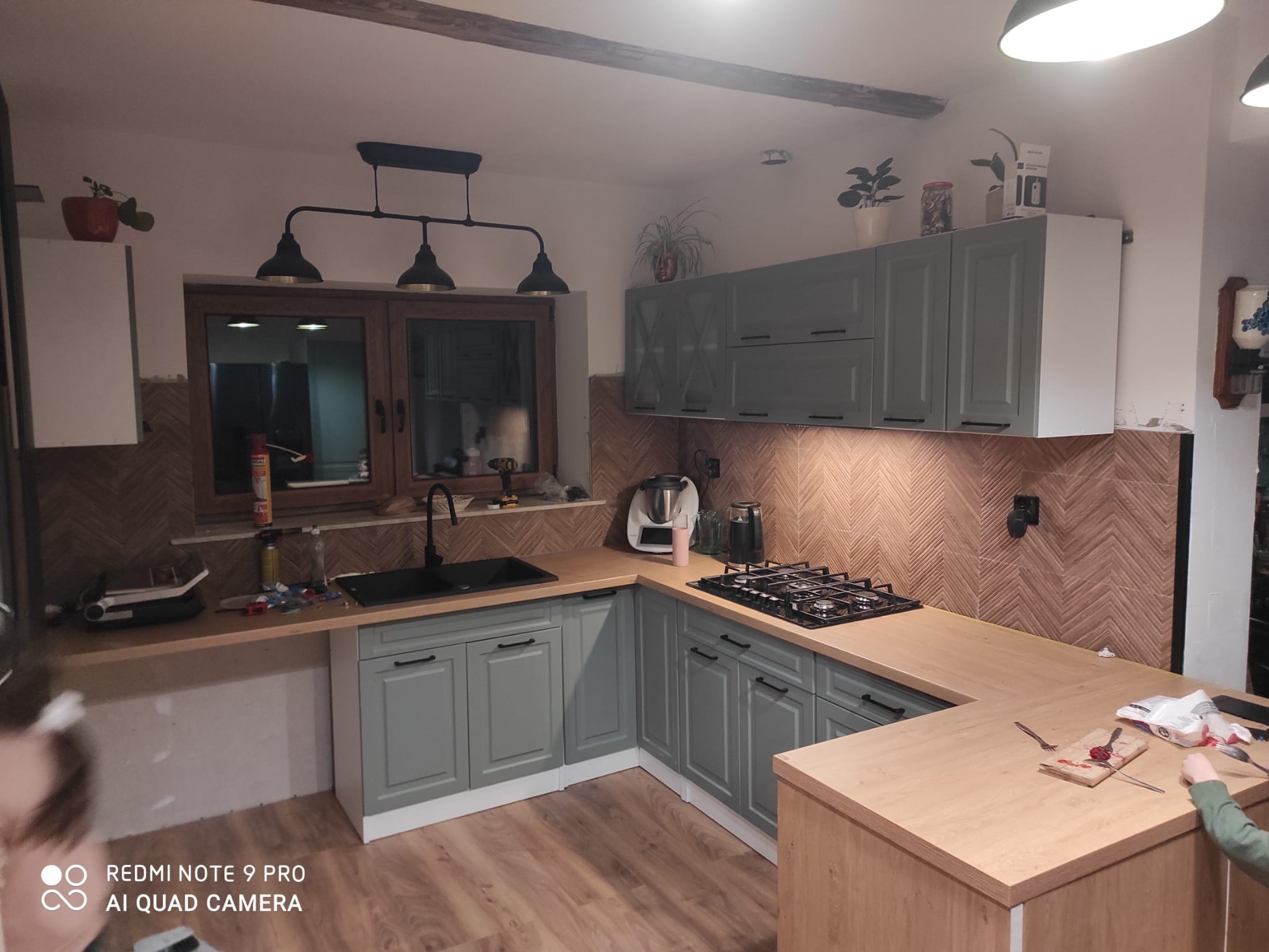 Kuchnia w stylu rustykalnym z szarymi szafkami, drewnianym blatem i oryginalną mozaiką na ścianie. Czarny zlew i lampa nad oknem dodają charakteru. Widoczna podłoga z paneli.