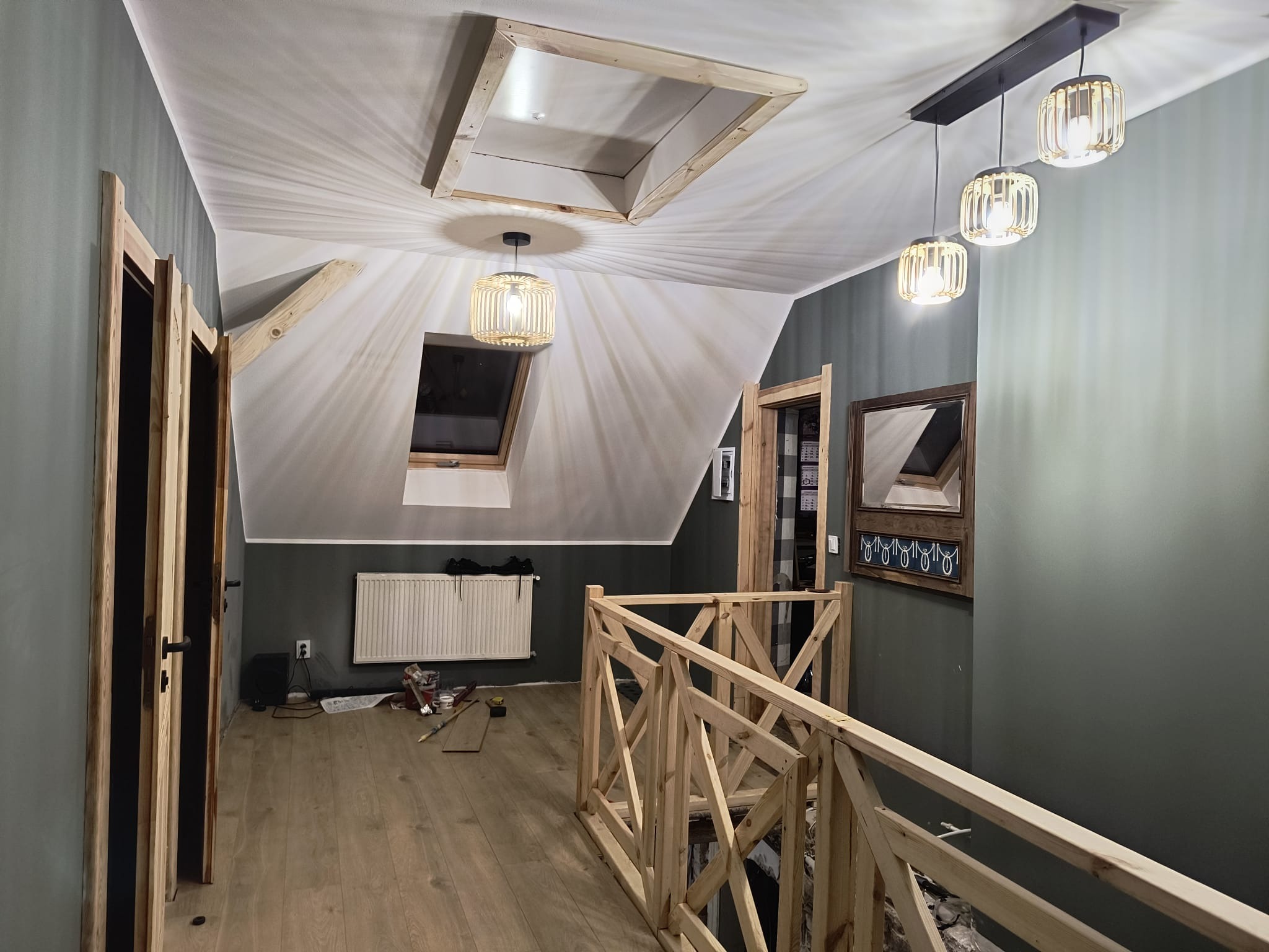 Poddasze w trakcie wykańczania: drewniana balustrada, obudowa okna dachowego i drzwi. Widoczne narzędzia i materiały budowlane. Oświetlenie sufitowe.