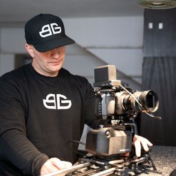 bartekgrzegorczyk.pl - Kamerzysta w czarnej bluzie i czapce z logo obsługuje profesjonalny sprzęt filmowy na statywie szynowym w jasnym wnętrzu. Gotowy do nagrywania.