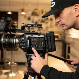 bartekgrzegorczyk.pl - Operator z czapką reguluje ustawienia profesjonalnej kamery Blackmagic na statywie, z obiektywem 35mm, w jasnym wnętrzu. Skupienie na ekranie kamery.