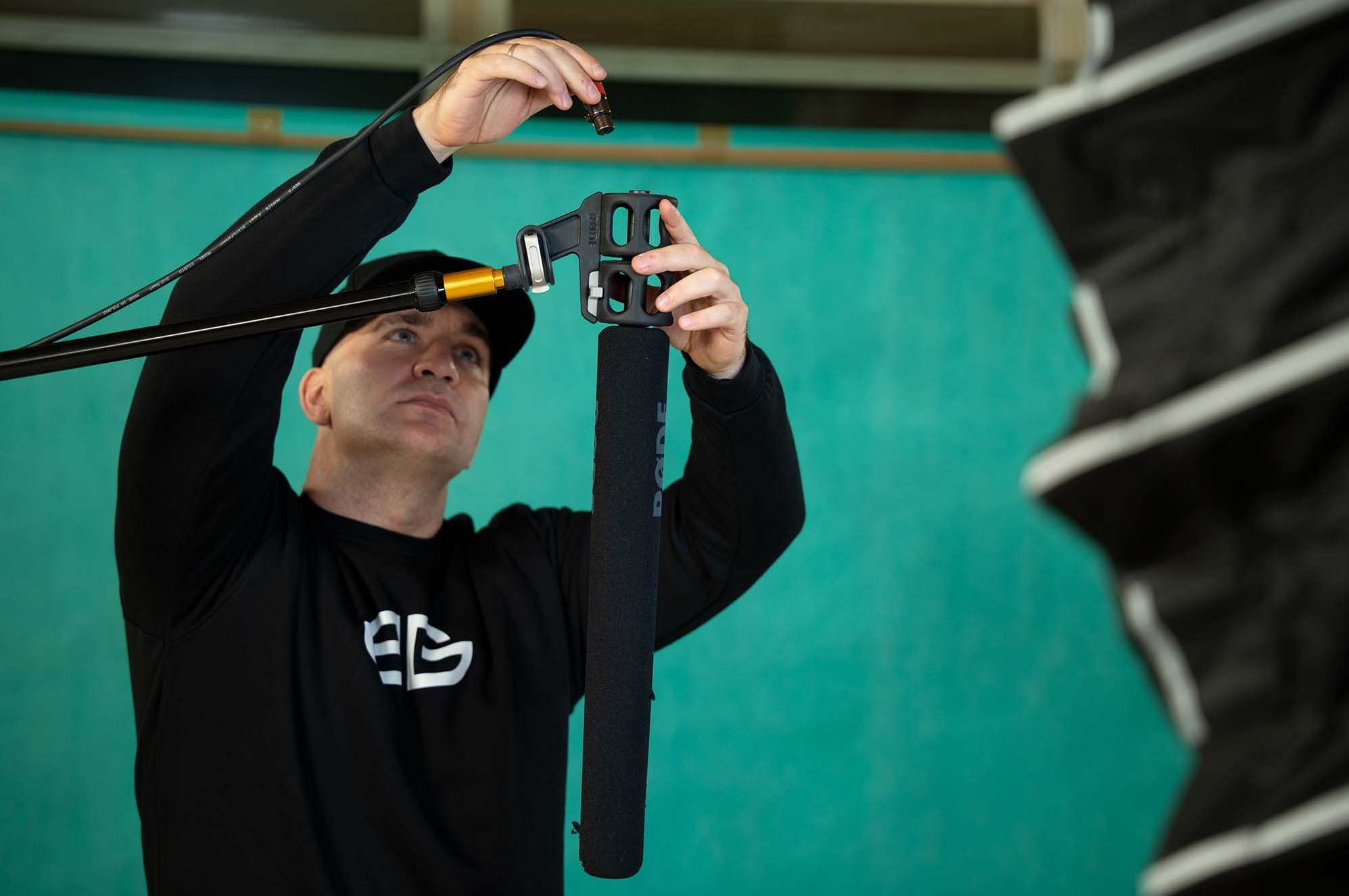 Mężczyzna w czarnej bluzie łączy mikrofon Rode z uchwytem na tle turkusowej ściany. Widoczny softbox po prawej stronie kadru. Ujęcie z dołu, skupione na sprzęcie.